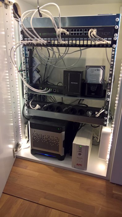 Serverskåp med nätverksutrustning, switch, NAS och HP server belyst av LED-lampor i ett överskåp.