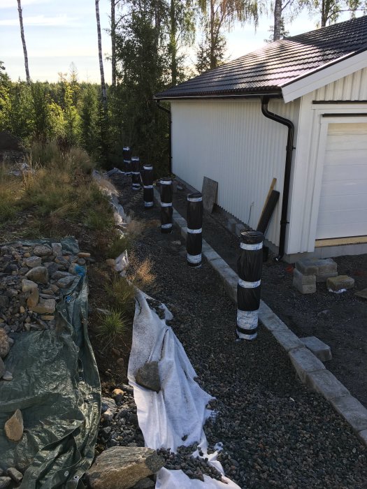 Plintar insvepta i svart plast med armeringsjärn, vid husfasad och grushögar, i byggprojekt.