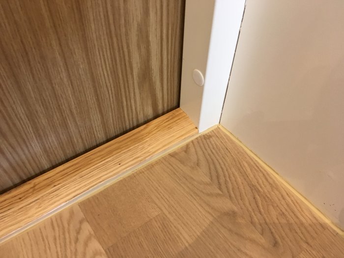 Ett hörn av ett rum med träimitation laminatgolv och en dörrtröskel fogad med Casco S40 i en ljus träfärg.