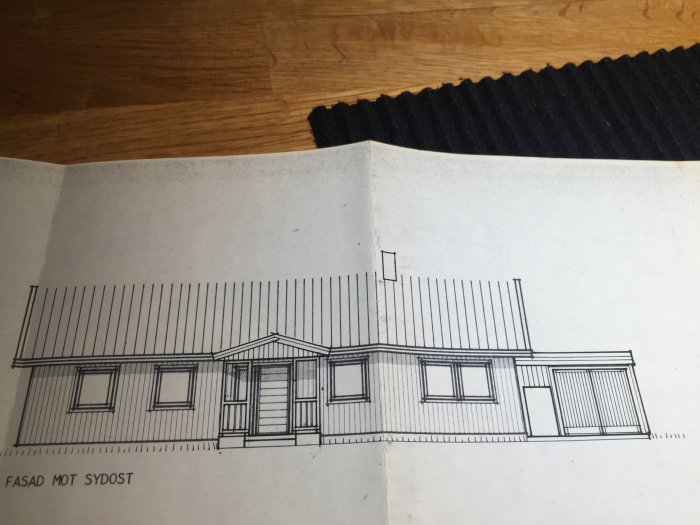 Ritning av enplanshusets fasad mot sydost på papper, visar husets design med fönster och dörr.