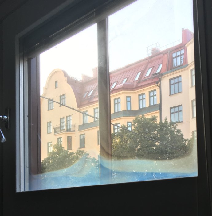Utsikt genom ett fönster med tecken på torkad kondens längs nedre kanten, skymmer delvis sikten till en gul byggnad.