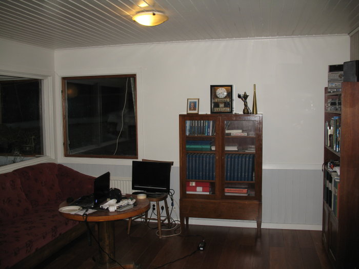 Renoverat vardagsrum med vitmålade väggar och bevarad bröstpanel under fönstret, möbler och hyllor med böcker.