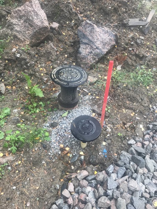 VA-lock i marken med inskription, bredvid en röd markör och omringat av grus och stenar.