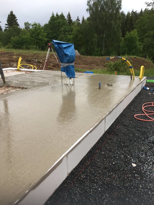 Nyligen gjuten betonggrund för hus med uppsatta formar och skyddad arbetsutrustning i regnigt väder.