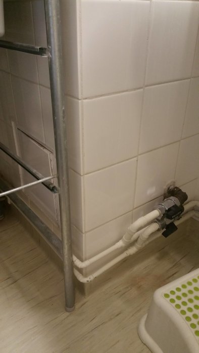 Badrum med synliga utanpåliggande rör och en ballofix nära en kaklad vägg och en fotpall.
