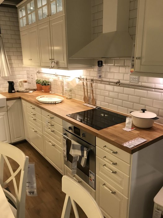 Vitt lantkök med trådbackar och trähantverk från IKEA, vita kakelväggar och moderna vitvaror.