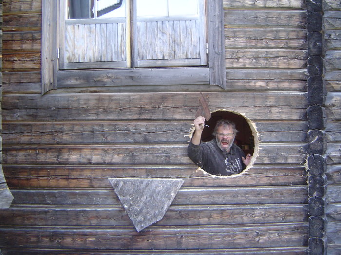 Man holding a piece of wood, shouting through a circular hole in a log cabin wall.