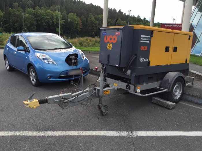 En blå elbil kopplad till en gul och blå dieselgenerator på parkeringsplats.