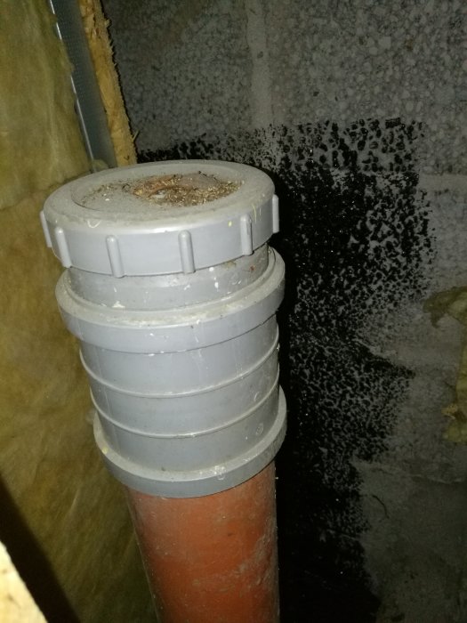Avloppsrör med grå vakuumventil i en källare, dolt bakom plywood.