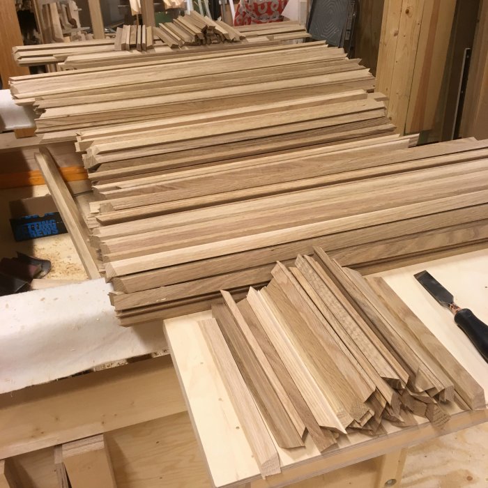 Staplar av slätade och kantbrutna glaslister i trä förberedda för montering, på en verkstadsbänk.