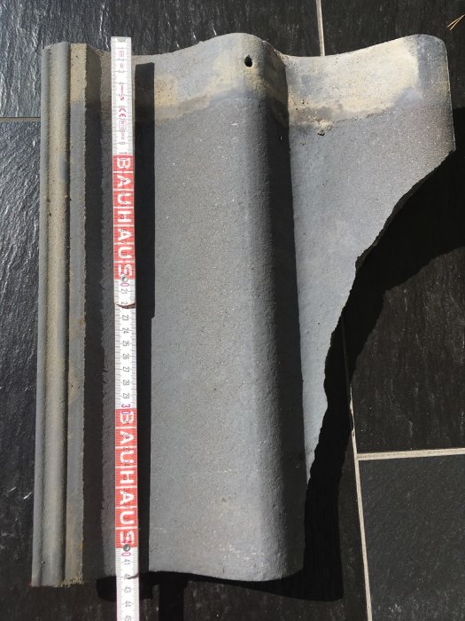 Söndrig betongtakpanna bredvid en meterstock på en svart yta.