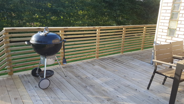 Nybyggd träveranda med klotgrill och utemöbler, omgärdad av ett trästaket.