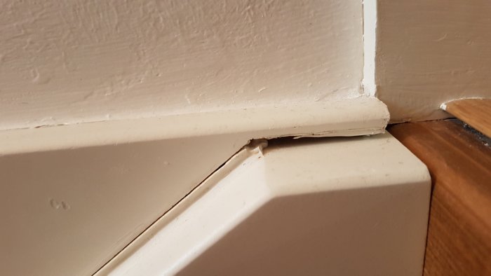 Spricka i hörnet där en vit vägg möter en vit taklist ovanför ett brunt trägolv.