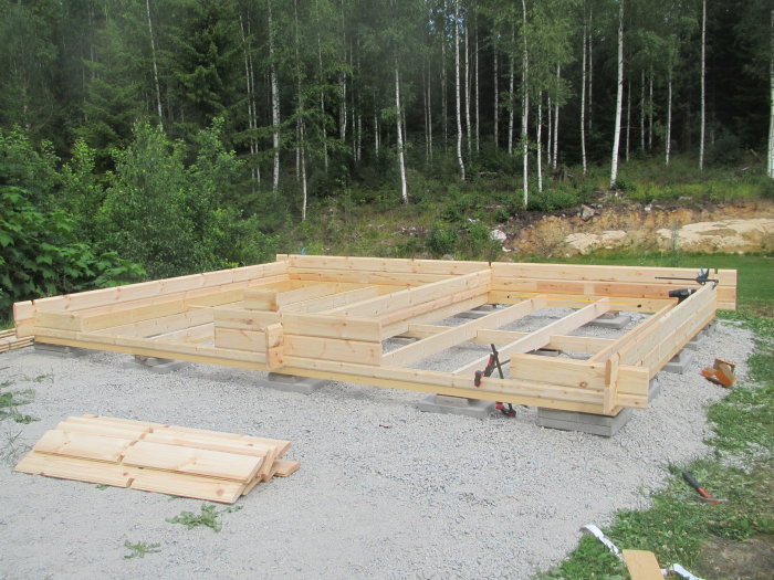 Början av bygget av ett vedbod och förråd med timmerprofiler på en grusunderlag, skog i bakgrunden.