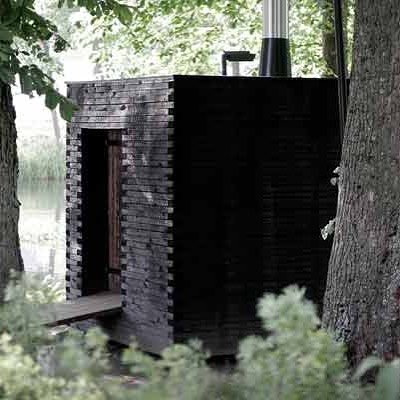 Svartmålad bastu i modern design, gömd bland träd vid vattnet, med synlig skorsten och entré.