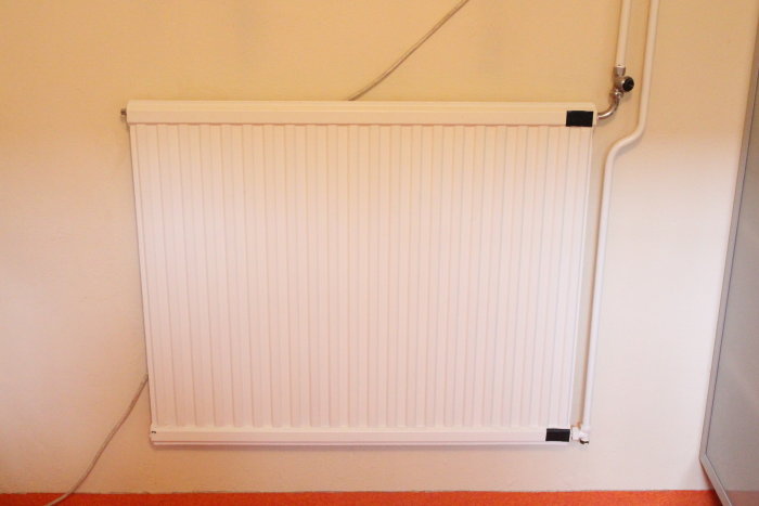 Vit panelradiator ansluten till väggen i ett rum, med synliga röranslutningar överst.