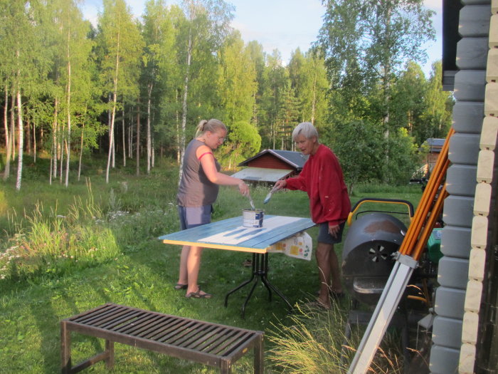 Två personer målar vindskivor och takfotsbrädor vid ett bygge i ett soligt landskap.