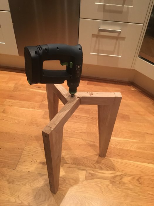 En handhållen borr placeras vertikalt på ett hemmagjort trästöd på ett parkettgolv.