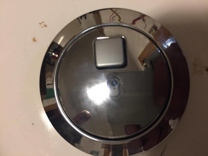 Reflektiv toalettspolknapp i krom med kvadratisk knapp och synlig mobiltelefon i spegling.