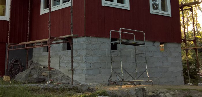 Oslagmurad lecagrund på tillbyggnad vid röd stuga, med byggnadsställning och material utspridda.