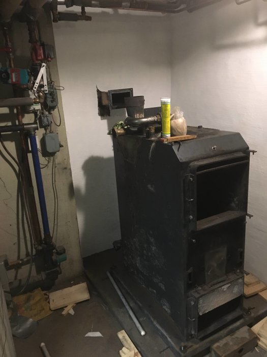En vedpanna med anslutet rör i ett källarrum med verktyg och rördelar på golvet.