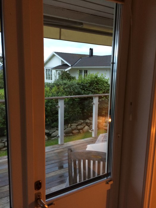 Vy genom öppen balkongdörr mot trädgård och vitt hus i skymningen.