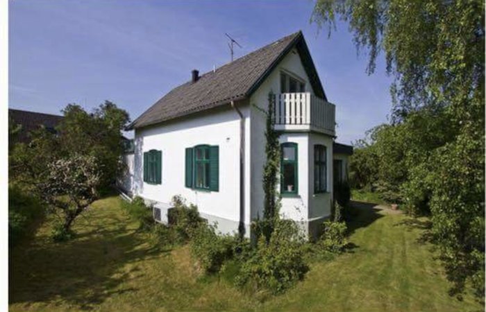 Vit villa med gröna fönsterluckor och veranda omgiven av en trädgård och fruktträd.