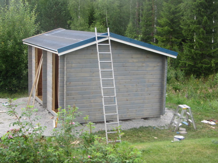 Nyfärgat grått förråd med öppen vedbod och stege lutad mot taket, omringat av natur.