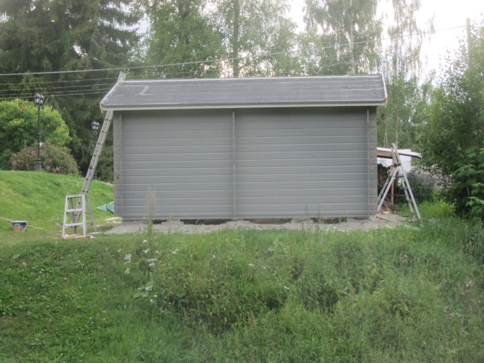 Nymålat grått förråd med nedfälld stege och målarutrustning intill en gräsmatta.