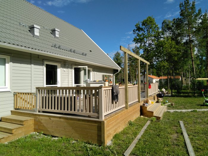 Nybyggd pergola intill ett grått hus med solsegel över, omgiven av en trädgård på sommaren.