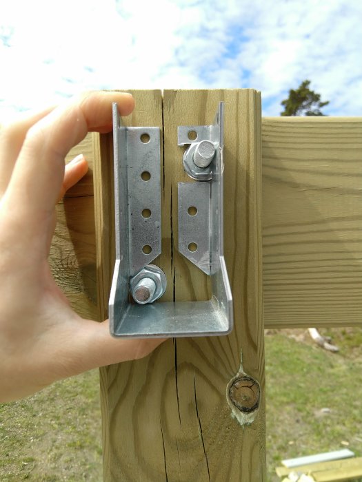 Hand installerar metallbeslag för pergolabygge mot en träpelare med blå himmel i bakgrunden.