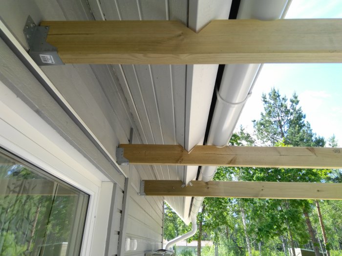 Obemålad träpergola under konstruktion monterad vid ett hus, med utsikt över träd och klar himmel.