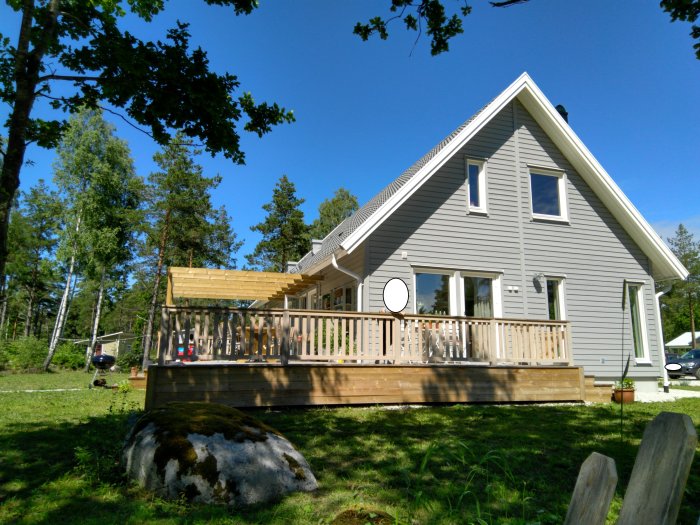 Nybyggd träpergola vid en gråmålad villa, omgiven av en trädgård med grönt gräs och träd.