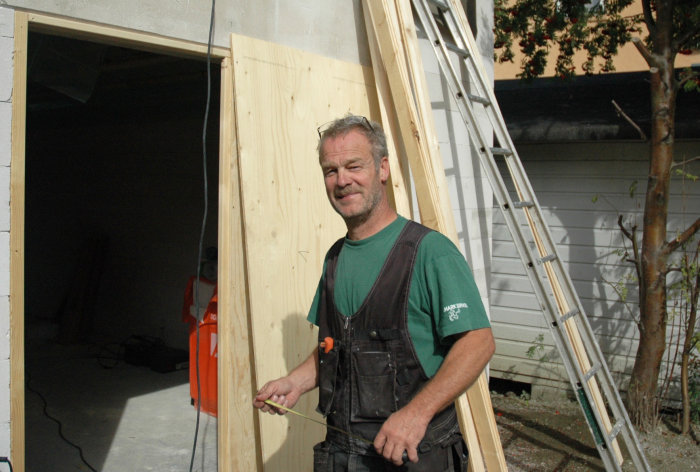 Leende snickare står framför en träkonstruktion med en måttband i handen.