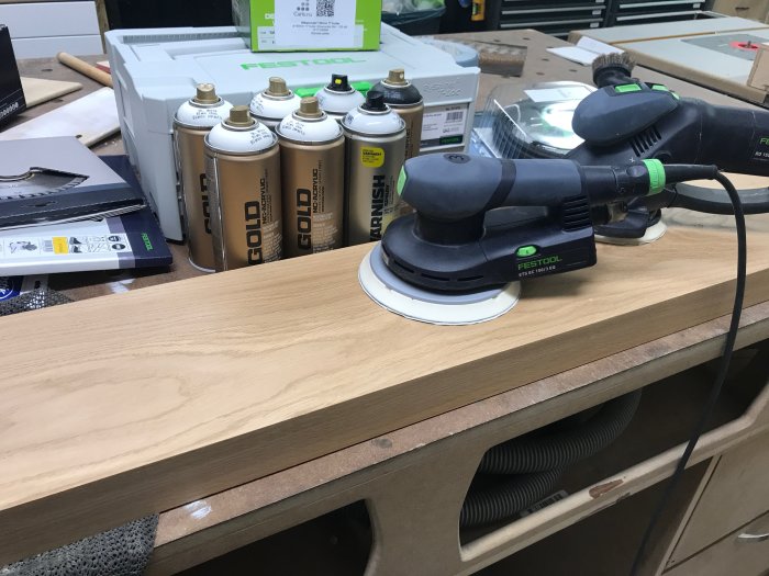 En Festool ETS 150/3 slipmaskin på en arbetsbänk bredvid förpackningar av Carls.nu slippapper och sprayburkar.