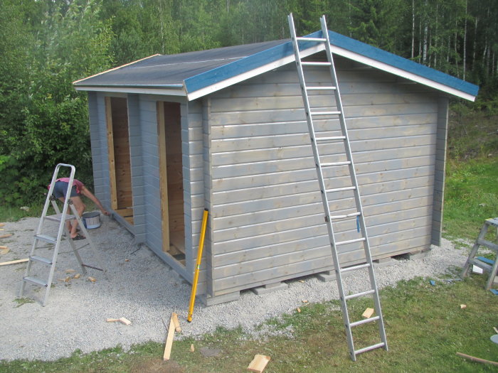 Person installerar dörr till vedbod, stege lutad mot vedbod i soligt väder.