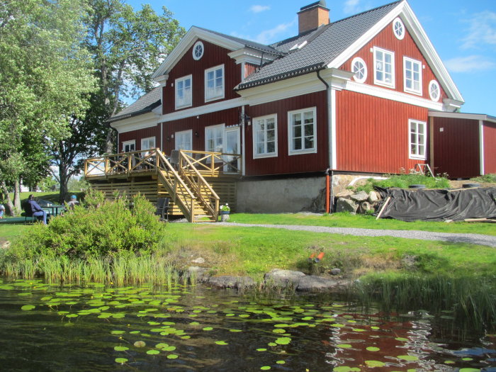 Röd trävilla med vita fönster och veranda vid en damm med näckrosor, under en klarblå himmel.