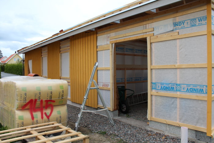 Byggprojekt med oisolerad del av fasaden, isoleringsmaterial, stegar och skottkärra.