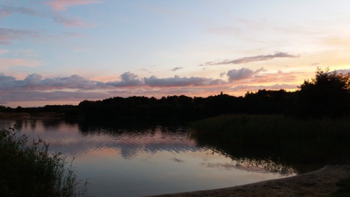 Solnedgång med färgglada moln speglade i en stilla sjö med sandstrand och träd i förgrunden.