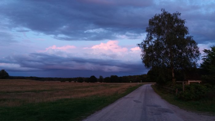 Landsväg vid kvällstid med färgstark himmel och moln, omgiven av fält och träd.