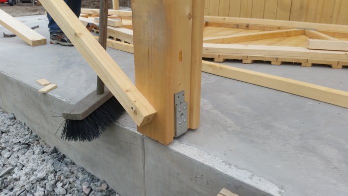 Trästolpe fastsatt i stolpsko på betongplatta med synliga borrade hål och ankarmassa.