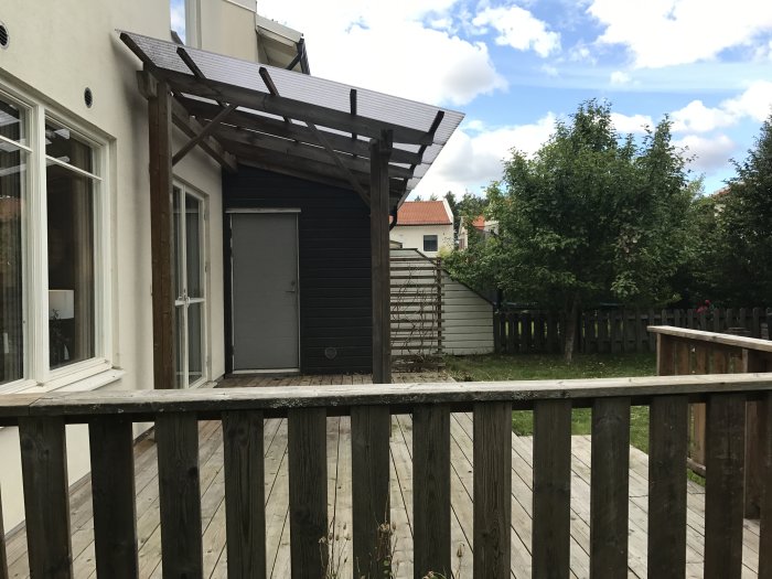 En befintlig trädgårdsterrass med pergola före renovering, omgiven av gräs och ett plankstaket.