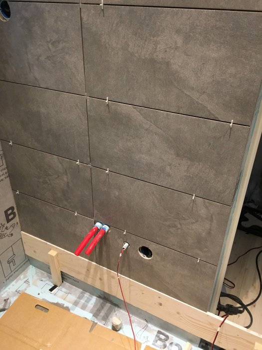 Ongoing tiling with large grey tiles in a bathroom, featuring tile spacers, and cutouts for plumbing with tools visible.