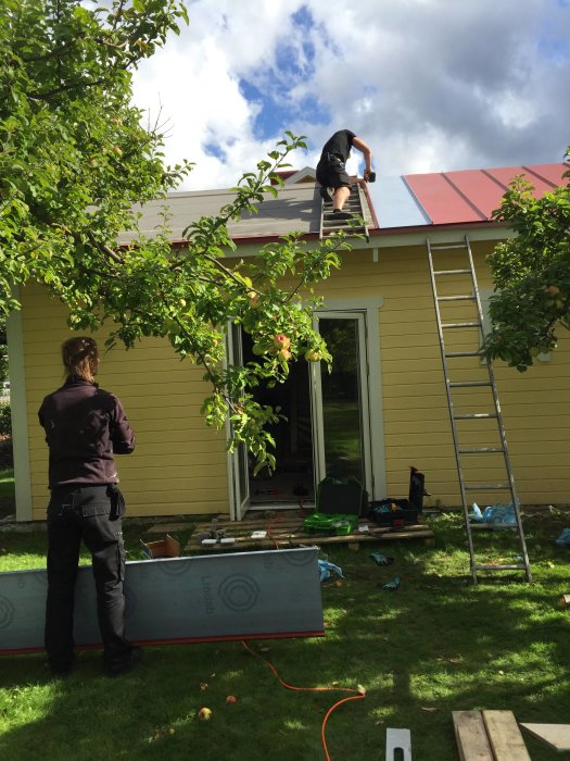 Två personer lägger rött plåttak på ett gult hus med gröna omgivningar och verktyg utspridda på marken.