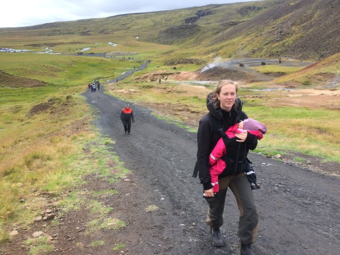 Person bär ett barn på en promenad i ett islandskt geotermiskt landskap med vandrare i bakgrunden.