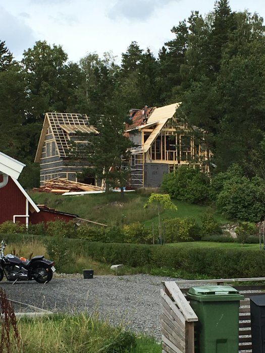 Hus under renovering med ojämn takkonstruktion och exponerad takstol mot en skogsbakgrund.