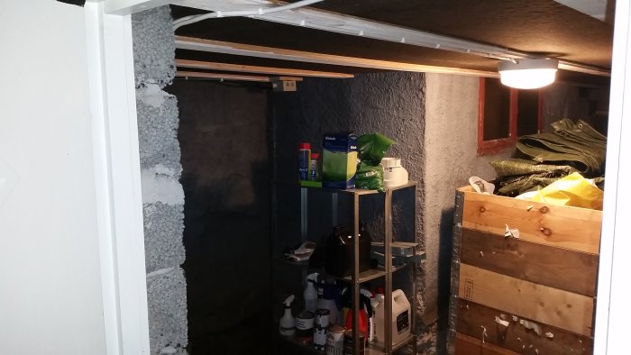 Källarförråd med hyllor, kablar och ändskydd på skena, isolering synlig på taket.