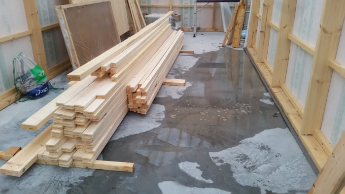 Vattensamling på en garageplatta med lutning inåt, omgiven av trämaterial och pågående byggnation.