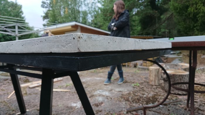 Böjd betongskiva på svart metallunderrede med synlig glapp mellan skiva och underrede, utomhus med person i bakgrunden.