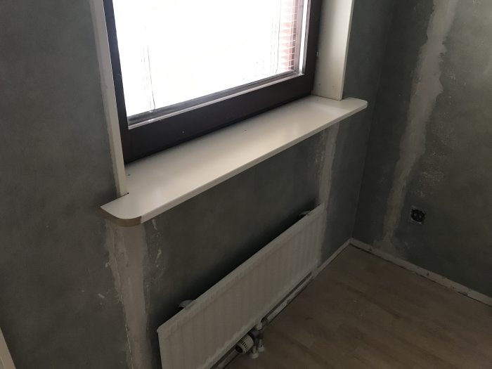 Ett rum under renovering med spacklade väggar och en nyligen installerad infälld fönsterbräda ovanför en radiator.